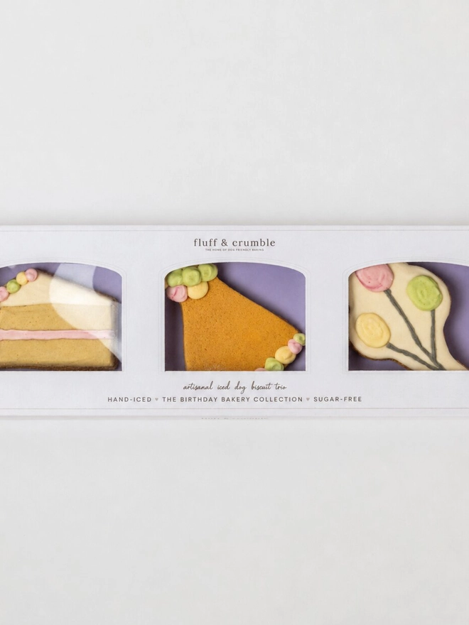 Boxed Celebration iced dog biscuit trio featuring a cake slice, party hat and balloon bone decorated in pastel yoghurt icing.