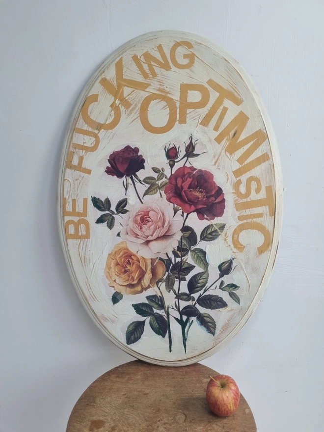 An art panel made from an upcycled table top. The image is vintage roses on a cream background with text that reads 'be fucking optimistic' in mustard yellow..