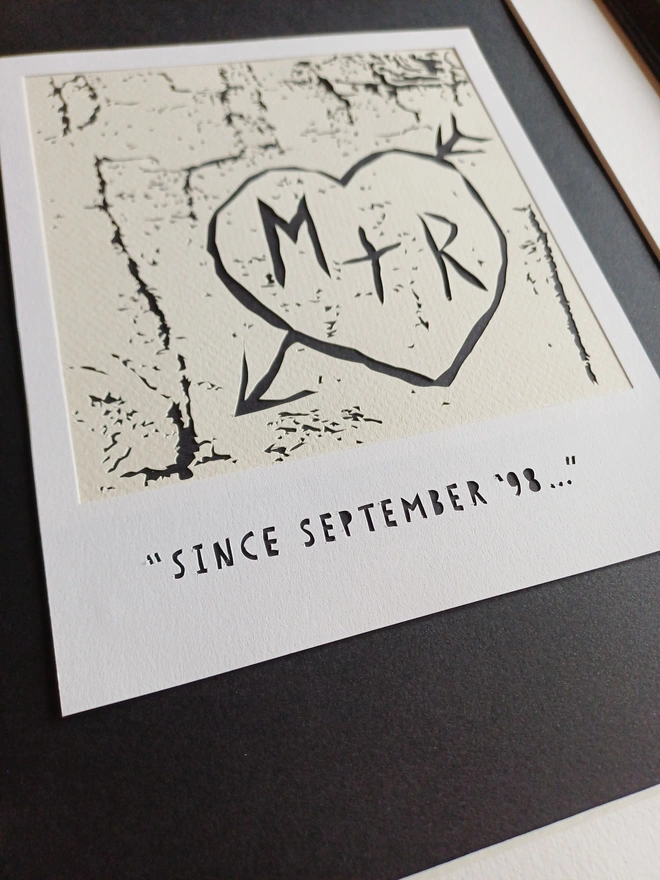 Papercut Showing A Polaroid "Photo" of Initials Carved Into Birch Tree Bark. Initials M + R Surrounded By Carved Love Heart. Paercut Text on Polaroid Frame Reads "Since September '98..."