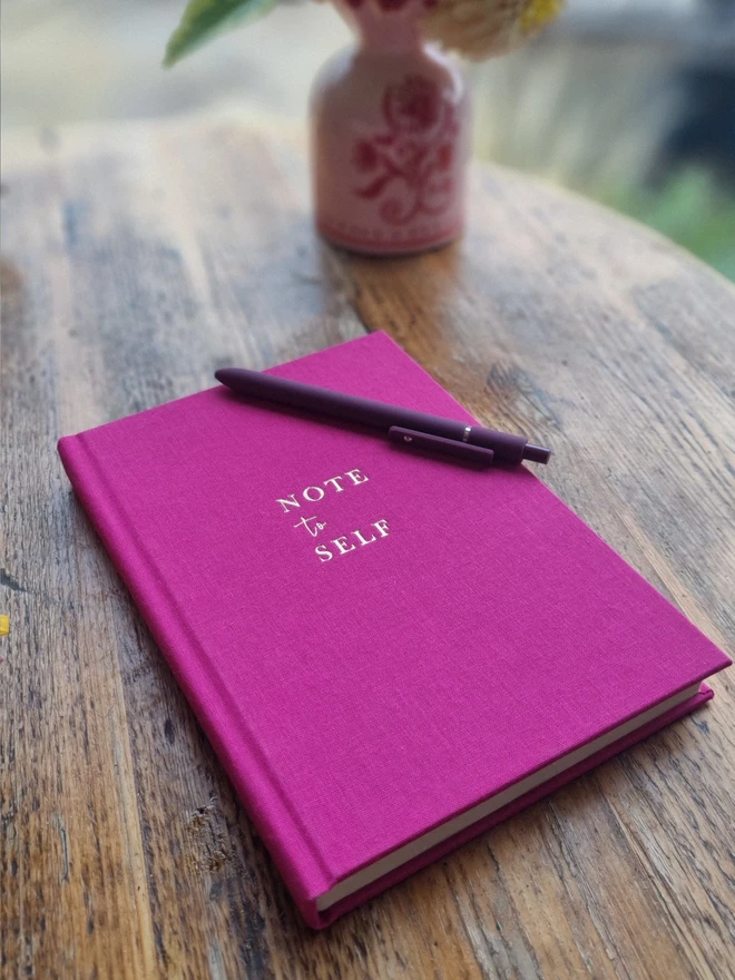 A bright pink notebook printed with the words 'Note to Self' in gold lettering rests on a wooden table top.