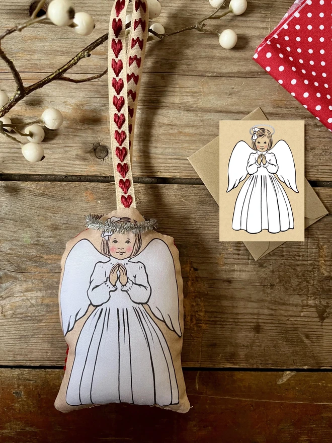 a photo of a plush angel christmas tree decoration next to a matching christmas card drawn in a vintage traditional childrens book style