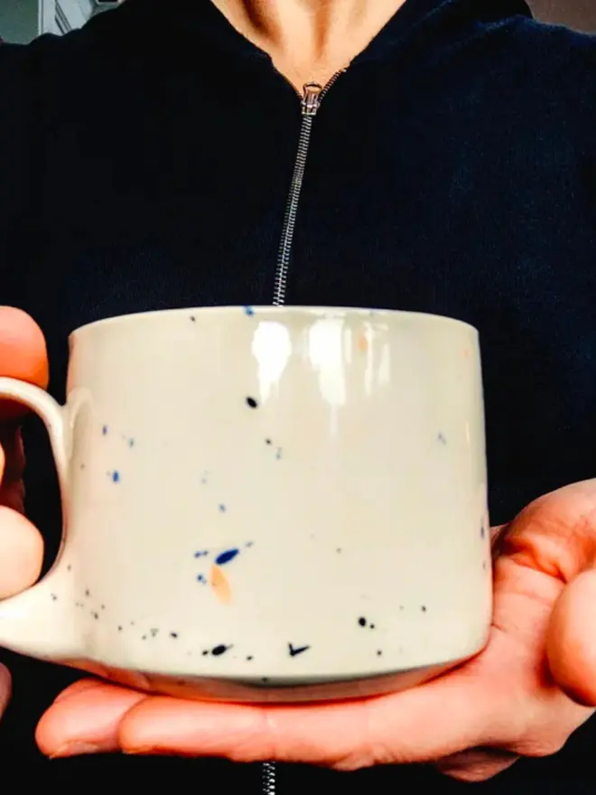 Small creamy white terrazzo cups with black, blue, and yellow splatters and glossy glaze being held by a person wearing a black top.