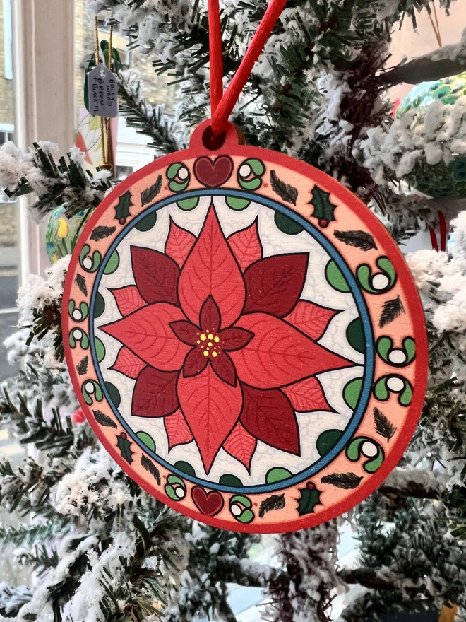 Multi coloured wooden tree decoration with a red poinsettia pattern at the centre. It is hanging by a red ribbon on a fake white christmas tree with other decorations. 