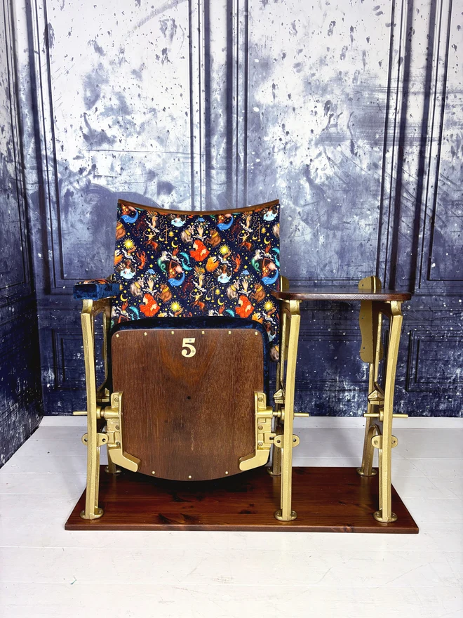 a vintage cinema seat with side table.  The seat is closed and features a wooden base with the number 5 on it.  The chair is upholstered in a navy velvet with astrology illustrations on it.  The chair is in front of a blue marbled wall. 