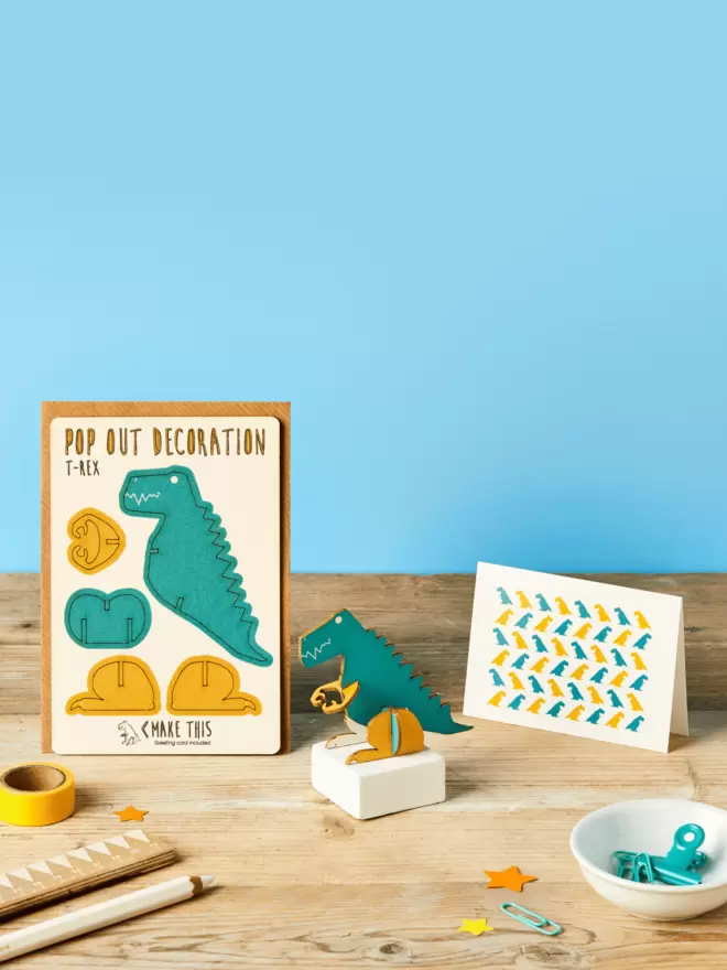 3D laser-cut dinosaur decoration and dinosaur pattern greeting card and brown kraft envelope on top of a wooden desk in front of a sky blue coloured background