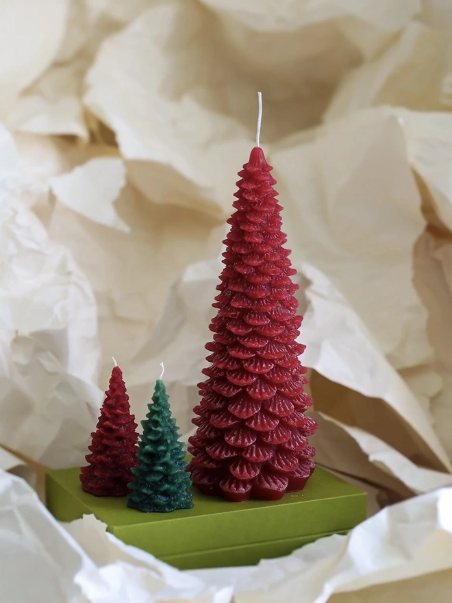 Large Christmas Tree Candle. a large deep red christmas tree shaped candle stands on a green box, nestled among crumpled cream tissue paper. a smaller red and a smaller dark green tree candle sit in front of it for scale, both with visible wicks. the textured, layered pine branch design and slightly variegated wax give all three candles a realistic, hand-poured finish.