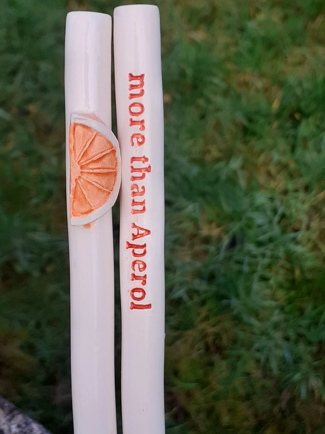 Handmade ceramic drinking straw set on a grassy background, featuring a raised orange slice decoration and a second straw with red text reading “more than Aperol.”