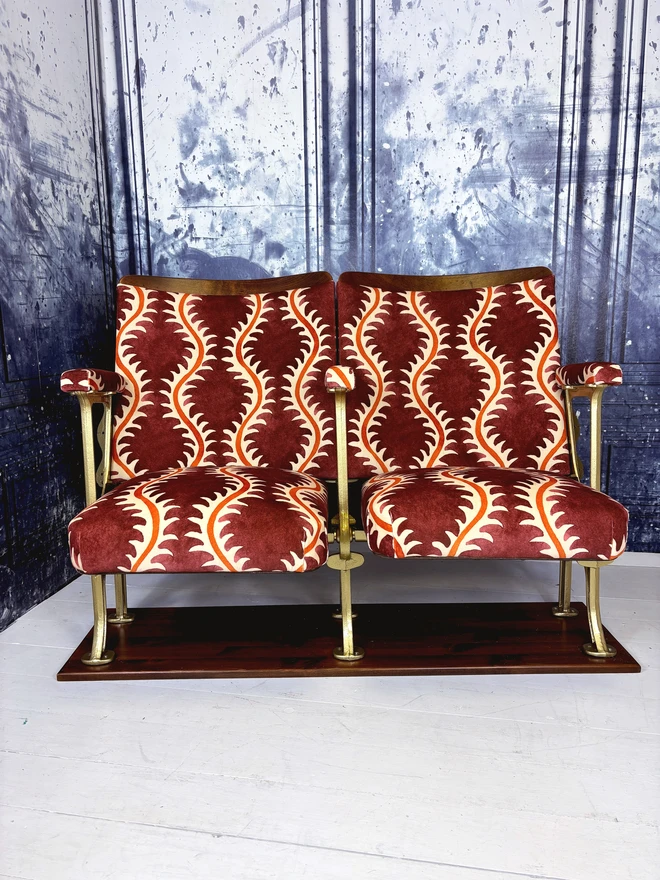 Set of two refurbished vintage cinema seats upholstered in 'Helter Skelter' redcurrant coloured velvet.  The flip down part of the seats are open.  The seats are mounted onto a wooden plinth in front of a blue marbled wall.