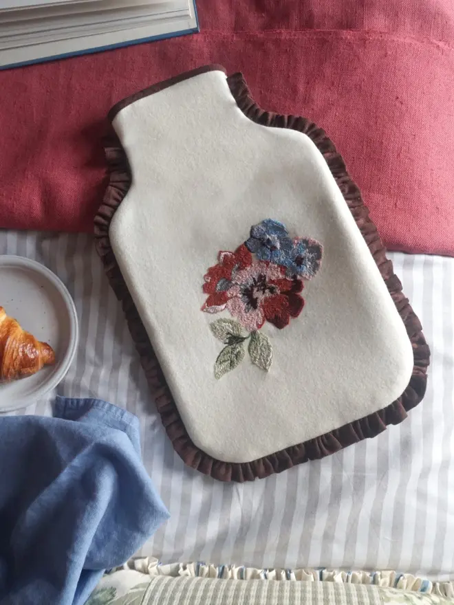 Embroidered Rose hot water bottle cover in cream wool with chocolate brown ruffle, styled with colourful bed linens.
