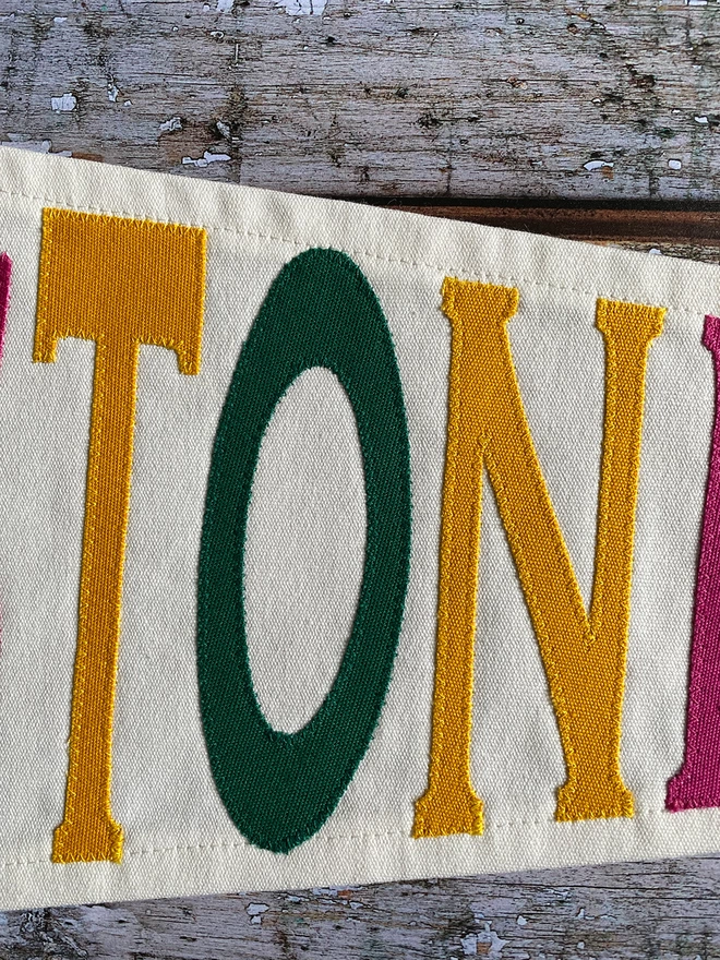 Detail of an ivory canvas Glastonbury pennant flag with the letter TON in yellow and green canvas.