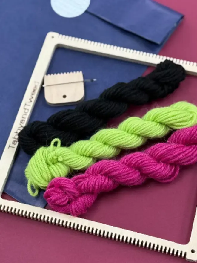 Square frame weaving loom kit with neon pink, green, and black tartan sample, yarn skeins, and wooden tools on a magenta background.