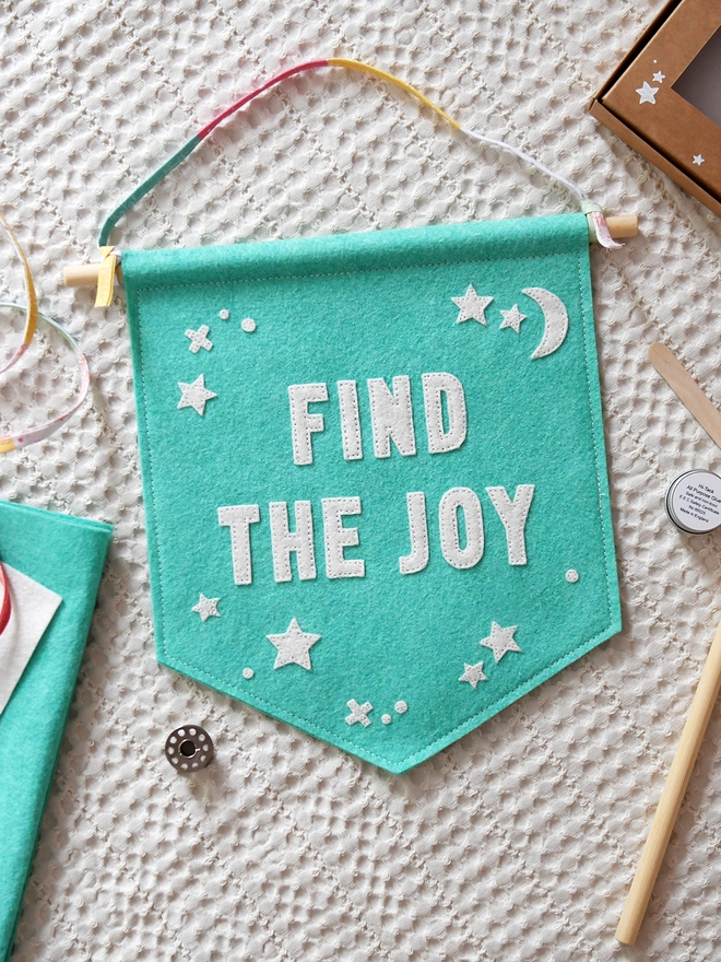 A turquoise felt wall hanging, with the words Find The Joy and white felt stars, lays on white fabric with various craft fabrics and items around it.