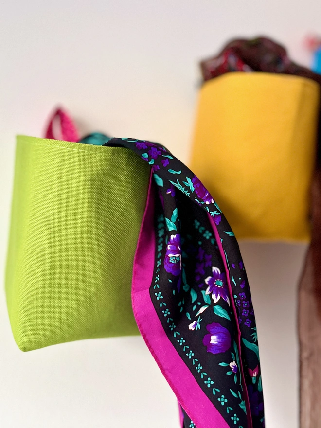 2 small fabric bags, 1 green, 1 yellow, hanging on a plain wall, holding colourful scarves.