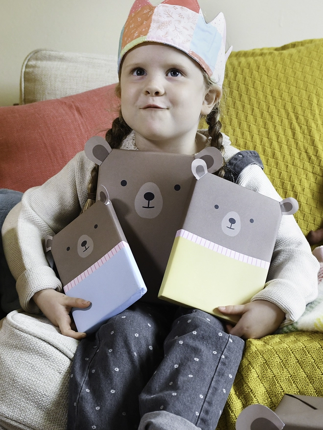 A young girl sitting on a sofa beside a Christmas Tree is holding three gifts wrapped up as brown bears.
