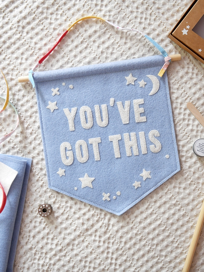 A blue felt wall hanging, with the words You’ve Got This and white felt stars, lays on white fabric with various craft fabrics and items around it.