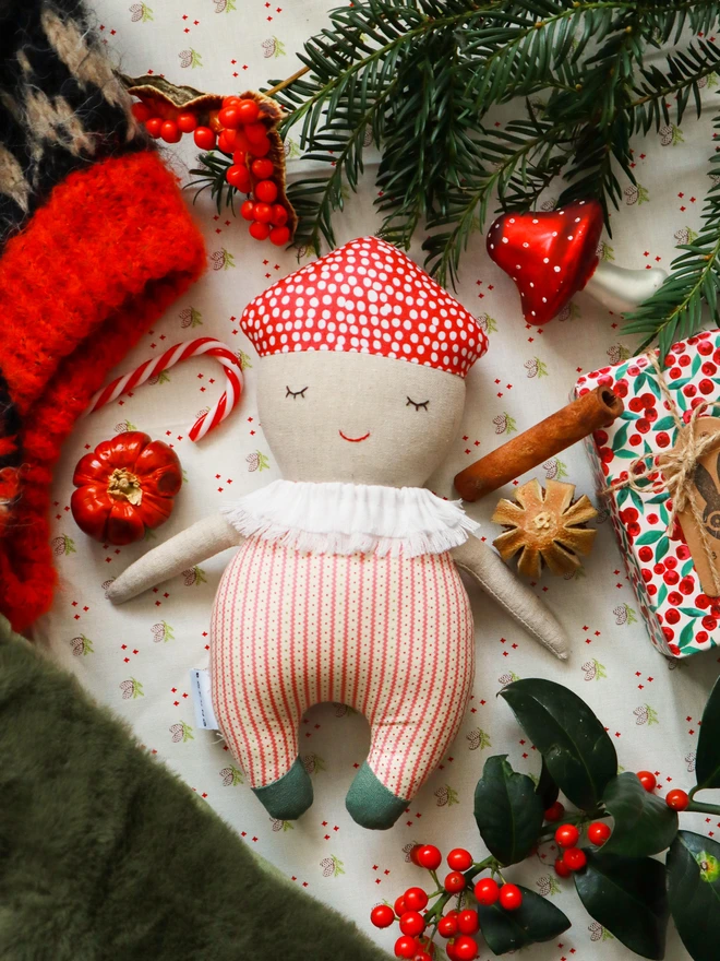 Soft handmade mushroom-themed doll with a red spotted cap, embroidered smiling face, ruffled white collar, red-striped body, and small green feet.
