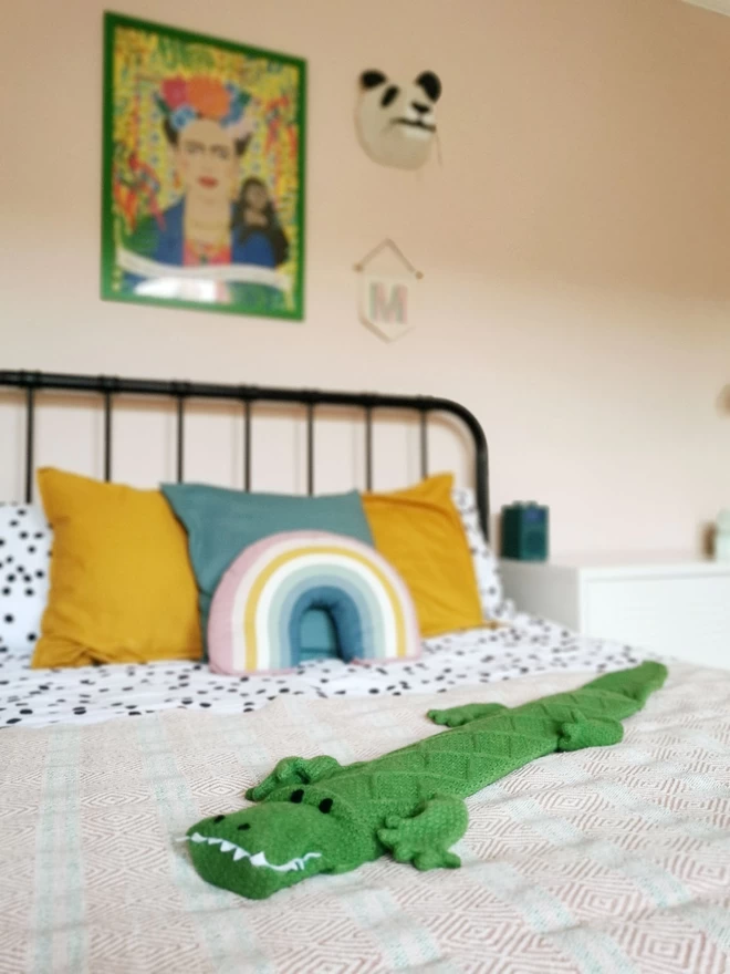 A bright green crocodile long hot water bottle is laid out on a double bed showing off his 1 metre length! Colourful rainbow cushions are propped up by the pillows and a picture of Frida Kahlo in a complimentary green frame hangs on the wall.