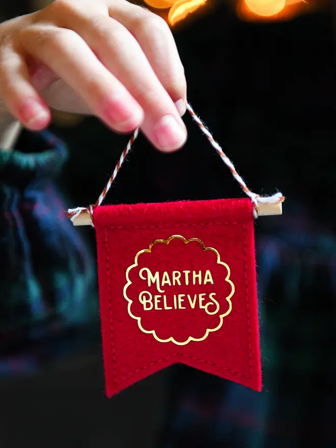 A child is holding a tiny red felt banner Christmas decoration with the words 'Martha Believes' in gold.