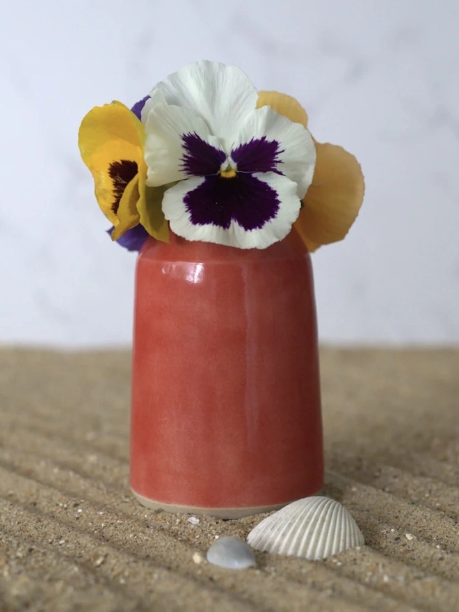 Classic Bud Vase, a colourful ceramic vase sitting on a plain surface against a grey backdrop. It is accompanied by some flowers and shells. 