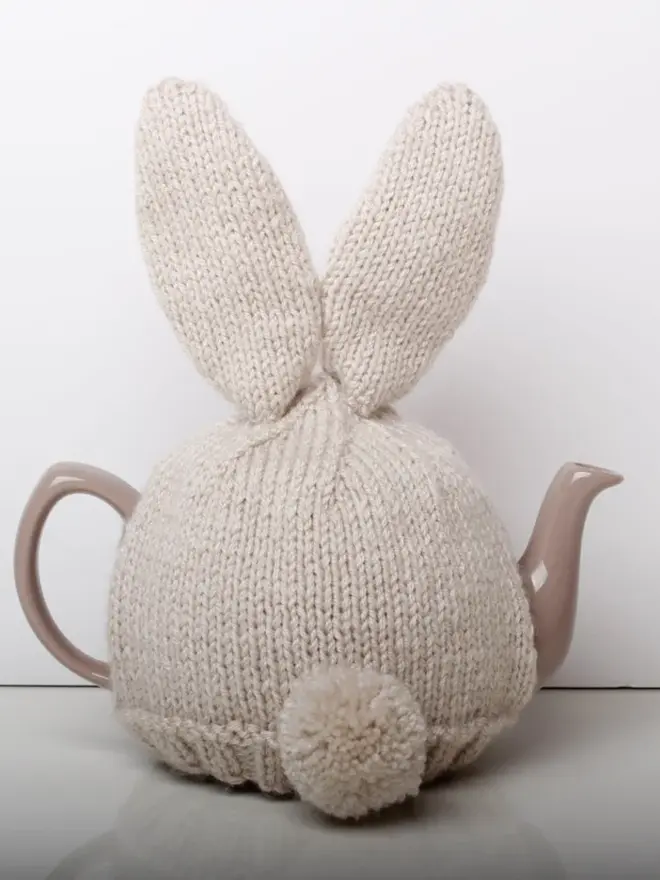 Hand Knitted Bunny Rabbit Tea Cosy, a rabbit bunny tea cosy sitting on a grey surface against a white backdrop. It fits over a white tea pot. 