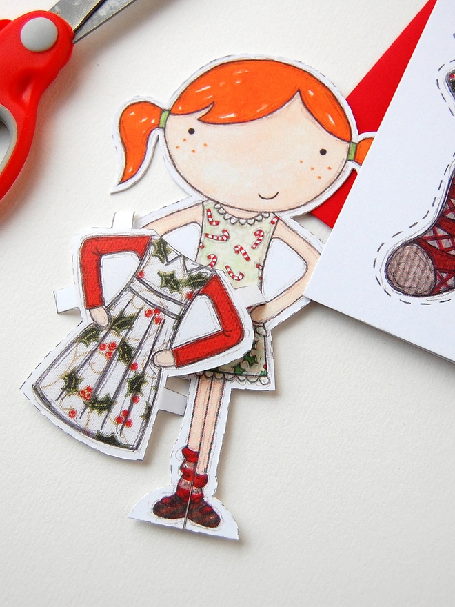 A Christmas card with an illustrated paper doll design is with a red envelope on a white desk.