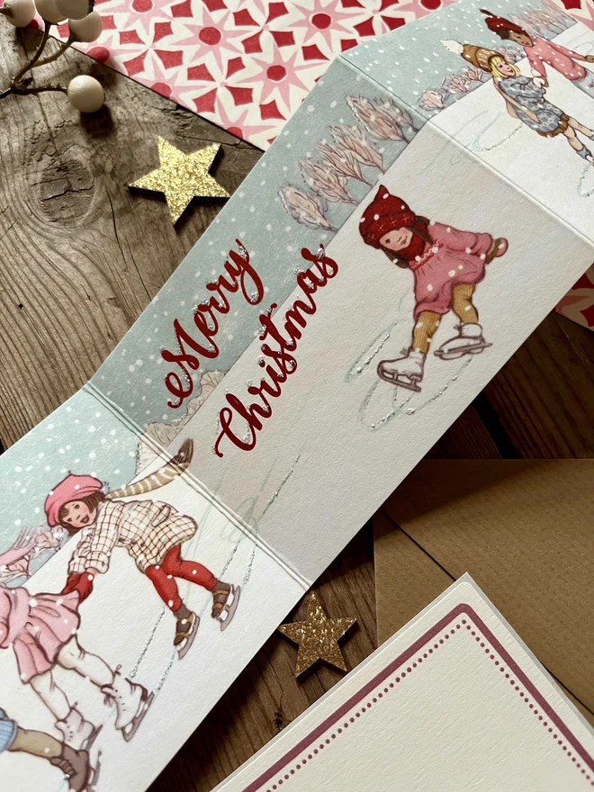a close up of an illustrated Concertina Christmas card featuring my storybook style illustration of children ice skating the card says Merry Christmas and has a retro vibe hand finished with glitter