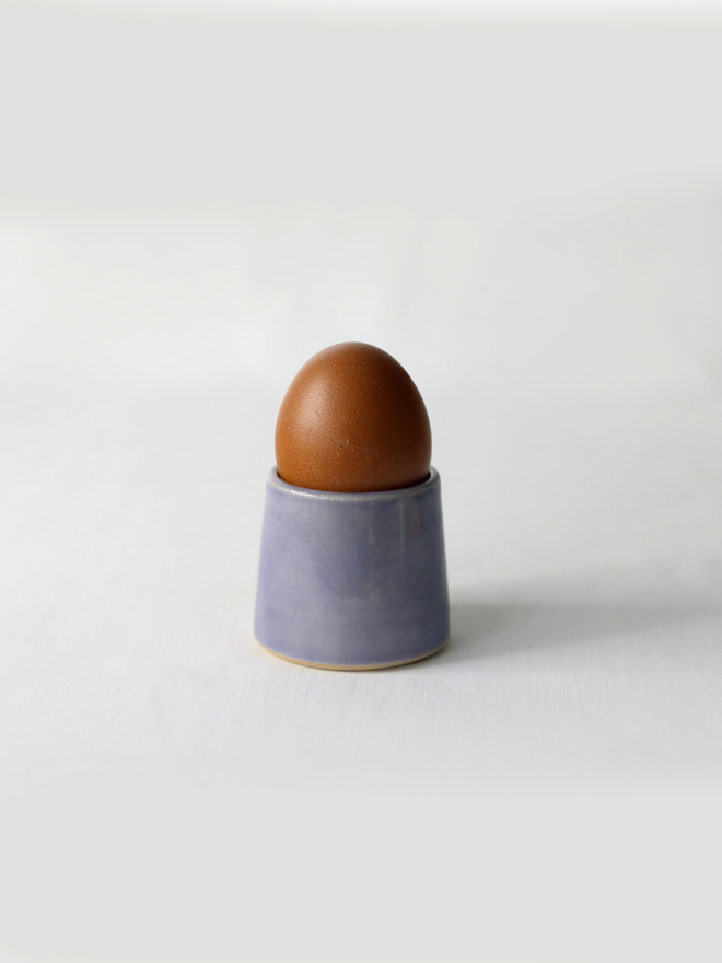 A handmade ceramic egg cup in amethyst purple glaze holding a brown egg, photographed against a plain white background