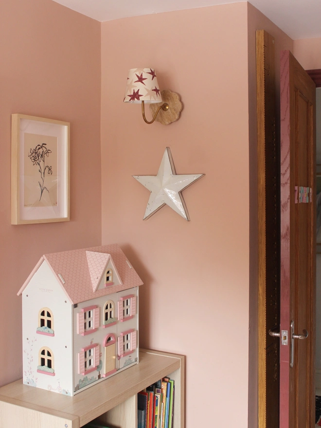 A wall-mounted empire light shade with bold red and pink stars, adding a playful accent to a girl's bedroom decor. The pink girls bedroom has a dolls house, books flower art. A star lampshade and colourful lampshade ideal for kids lampshade styling.