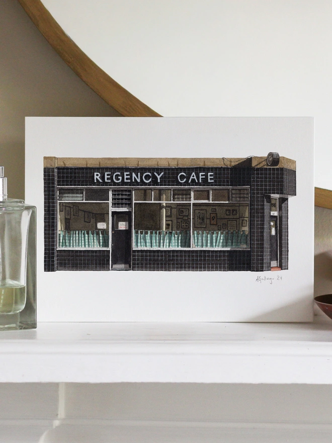 An architectural illustration of the Regency Cafe storefront on a print, displayed on a white shelf next to a perfume bottle and a small decorative bowl.