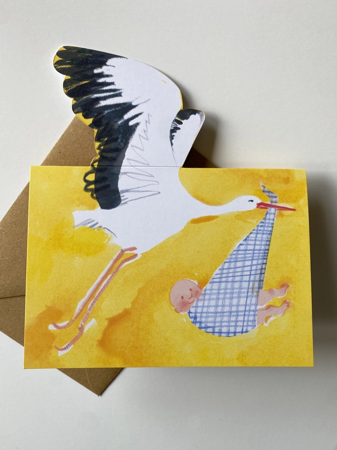 A new baby card by Esther Kent showing a black and white stork carrying a baby in a blue checked cloth, on a yellow background