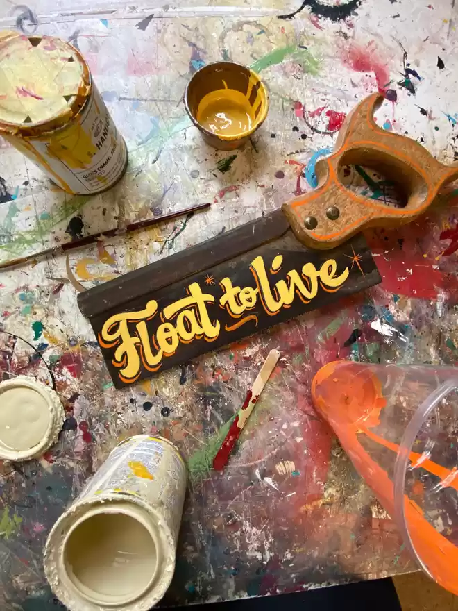Hand-painted vintage saw spelling 'float to live' in yellow and orange paint with paint pots and brushes surrounding it on a painty table.