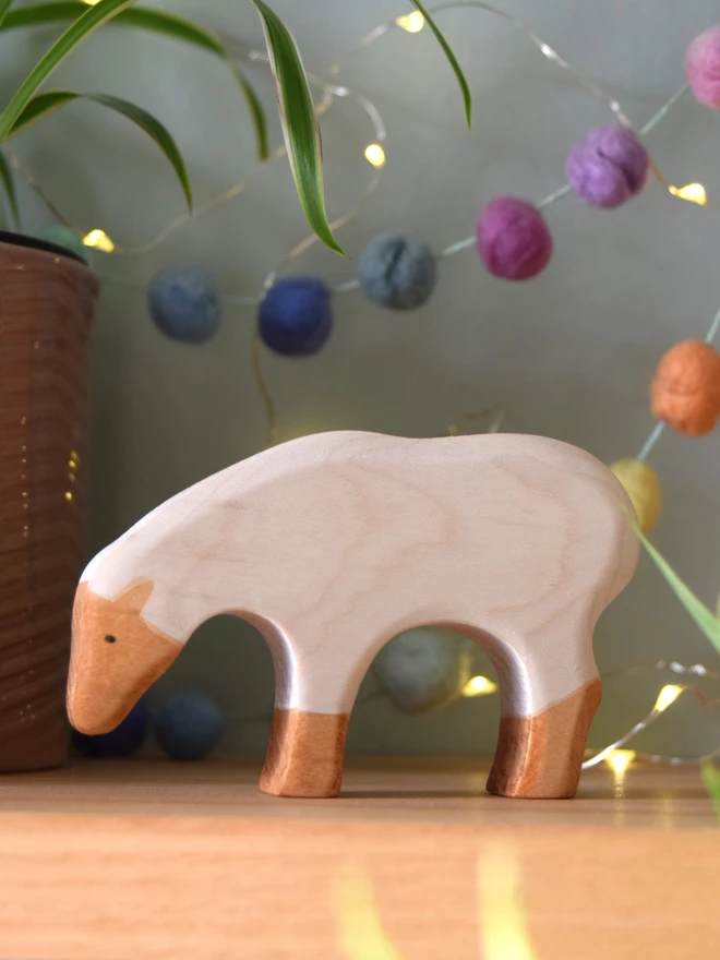 wooden toy grazing sheep in white on shelf with lights, plant and pom poms behind