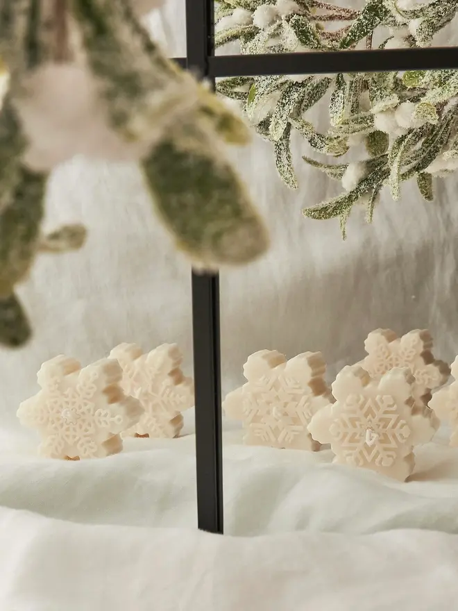 Classic Cream Snowflake Sculpture Candle. A row of cream snowflake candles standing upright on white linen beneath frosted mistletoe and foliage. Each candle has its own detailed snowflake design, so they make charming stocking fillers or festive table favours.