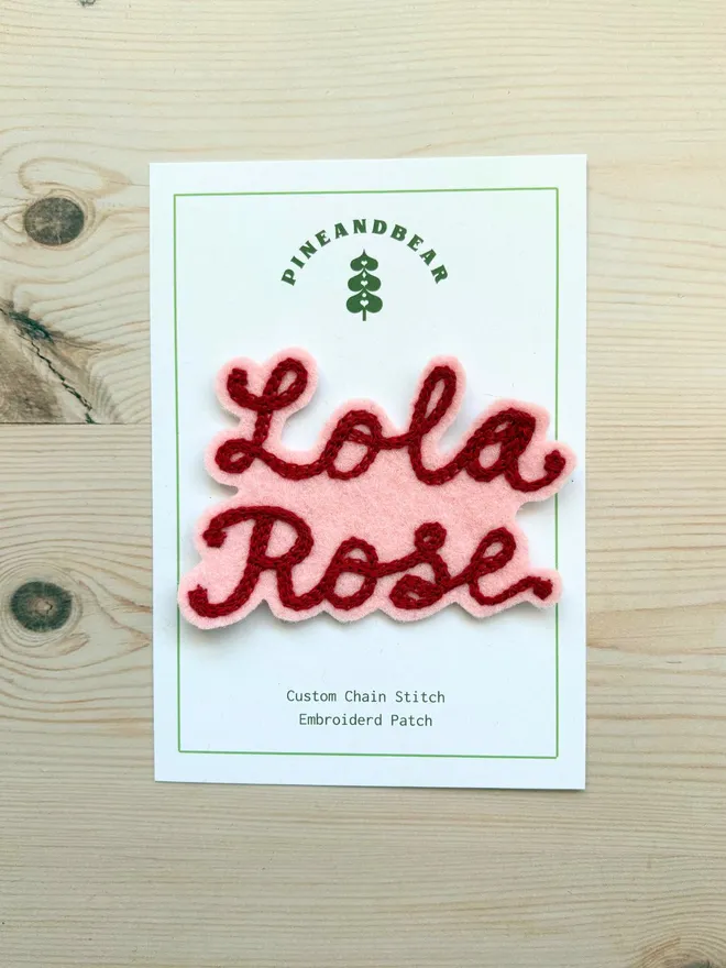 A chain stitch embroidered patch in pink and red spelling Lola Rose on a white presentation card