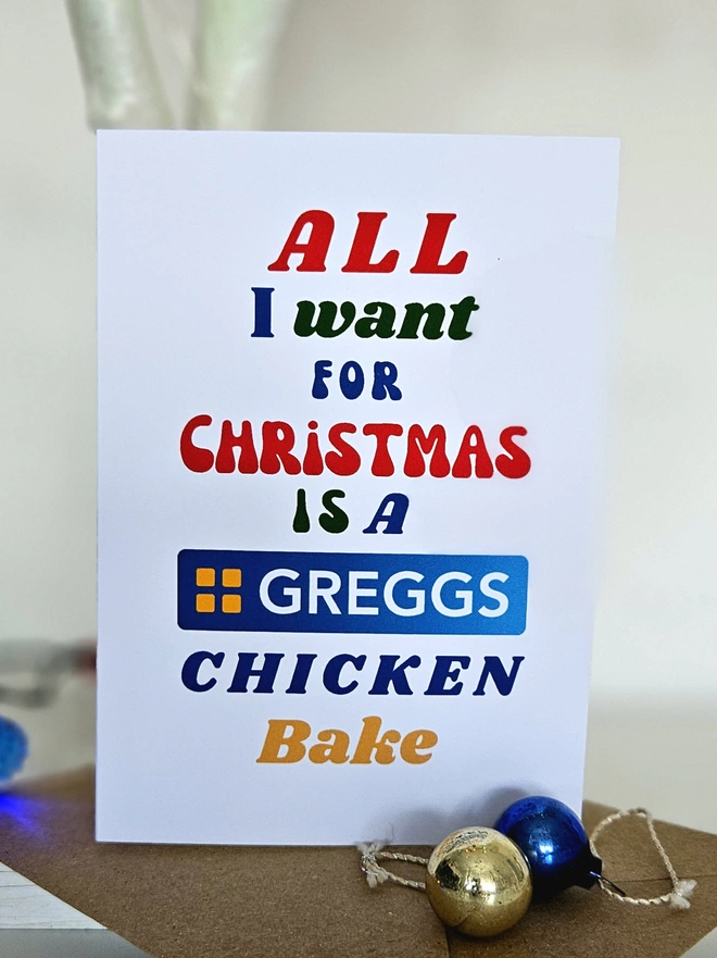 A colourful card Christmas Card that says All I want for Christmas is a Greggs Chicken Bake in retro style fonts on a white background.