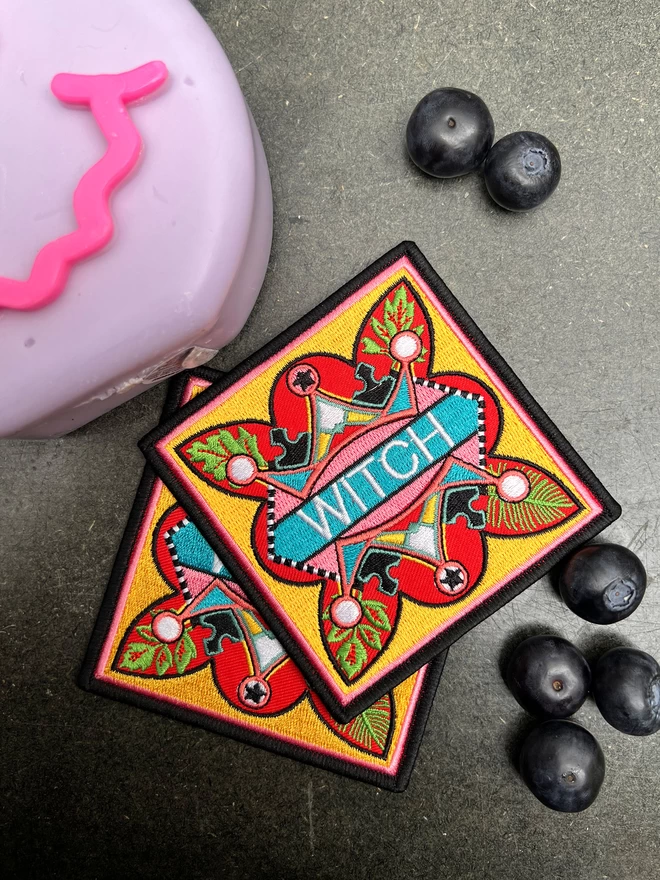 The patch sits on another patch, next to scattered blueberries and a pink plastic box. At the centre of the patch is the word “Witch” embroidered in white on a light blue strip. Surrounding this is a colourful, bold design which sits on a yellow background, with a thin pink inner border and a thick black outer border.
