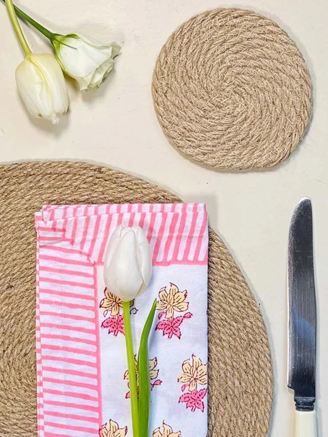 Close up of a round woven seagrass neutral placemats, with a flowery napkin on top shown on a light surface. 