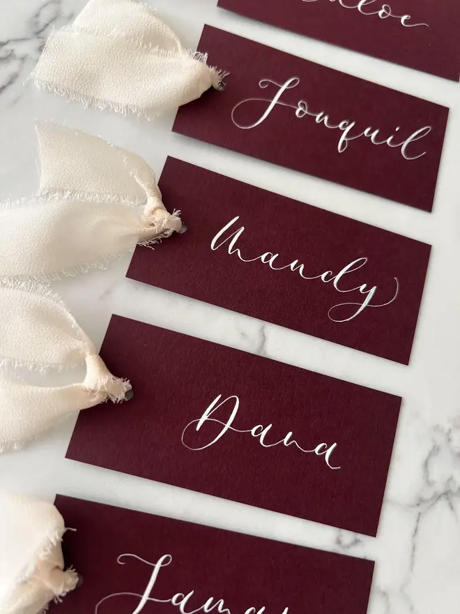 Personalised Calligraphy Wedding Burgundy Place Cards, burgundy place cards set together on a wooden table with champagne ribbons. 