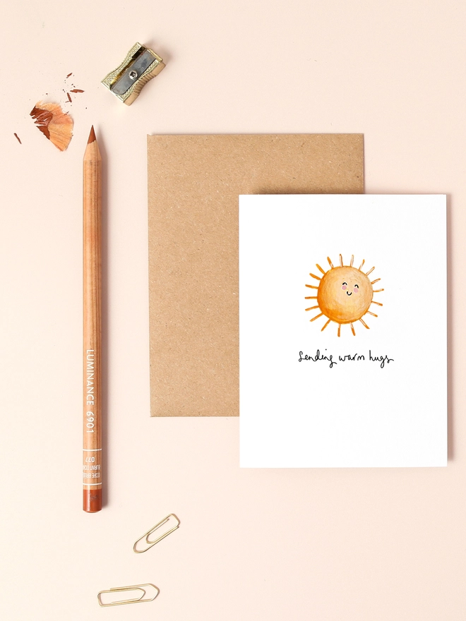 An image of a mini, rectangular white greetings card with an illustration of a smiley sun with a hand written 'sending warm hugs' message in black ink seen under the central illustration. The card is placed on top of a brown rectangular kraft envelope.