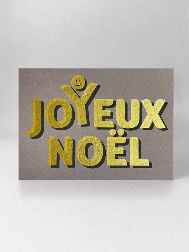 Golden text screenprinted onto grey card stock, spells out Joyeux Noel with a happy face above the Y, as though celebrating. Landscape card stood on a grey background, viewed front on.