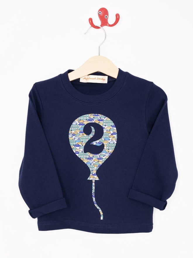 A navy cotton long sleeve t-shirt. The t-shirt features a balloon with the number 2 cut out from it, appliquéd in a cars Liberty print. Hanging on a hanger.