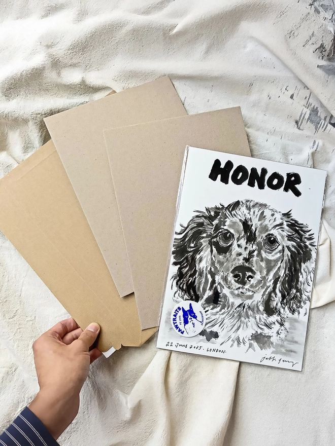 A table showing a personalised pet portrait by artist 'Dogs by Jeff' with a hard-backed envelope next to it on a table, held by a hand