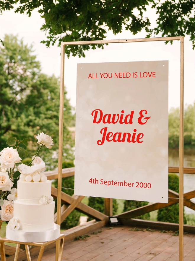 a Gold frame with a white board hanging from it, with the wording all you need it love, David and Jeanie and the date of the wedding in red lettering. the stand is surronded by a wedding cake and flowers in white and tree foliage