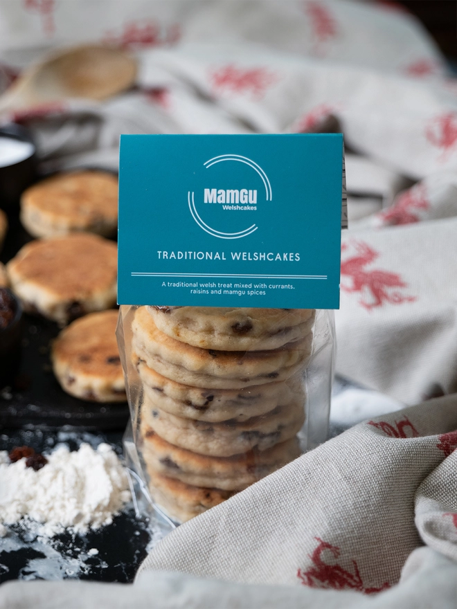 Packaged Traditional Welsh Cakes on a welsh dragon table cloth next to a griddle with cooking welsh cakes and flour