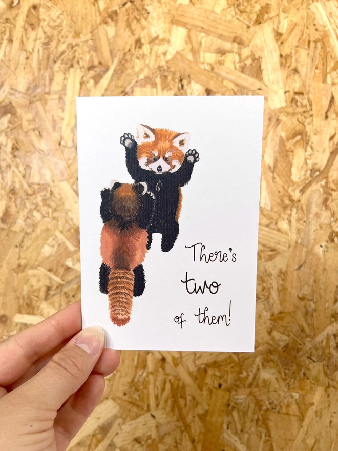 A greetings card featuring two baby red pandas stood up looking at each other with their arms in the air, next to the phrase “There’s two of them!”