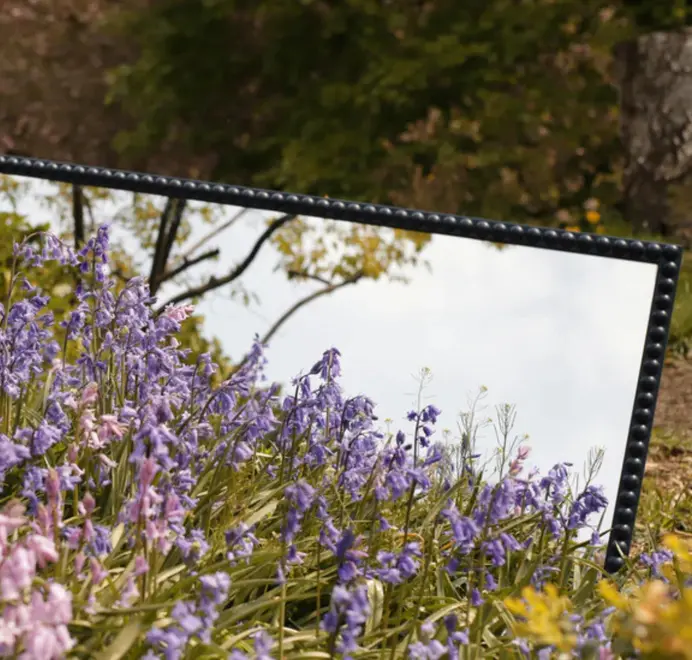 Floor-length bobbin mirror with a turned wooden frame standing among tall grass outdoors.