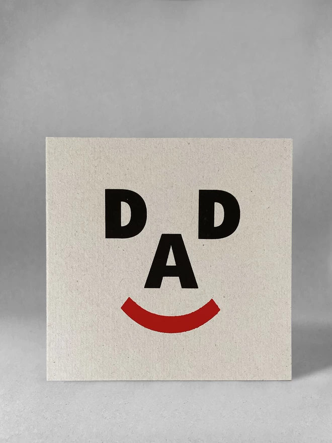  The letters D, A, D and a closed bracket symbol make up a happy face, screenprinted on recycled grey card. Stood front on, in a light grey studio set