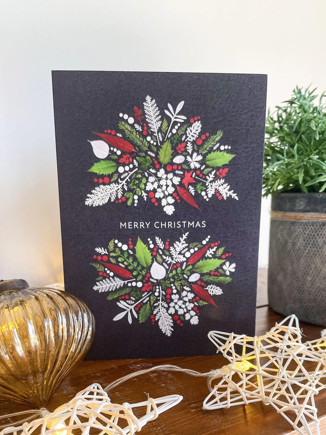 Christmas card with pressed leaf crown design - Holly and Ivy. On wooden bookcase with vintage glass bauble, star lights, potted plant
