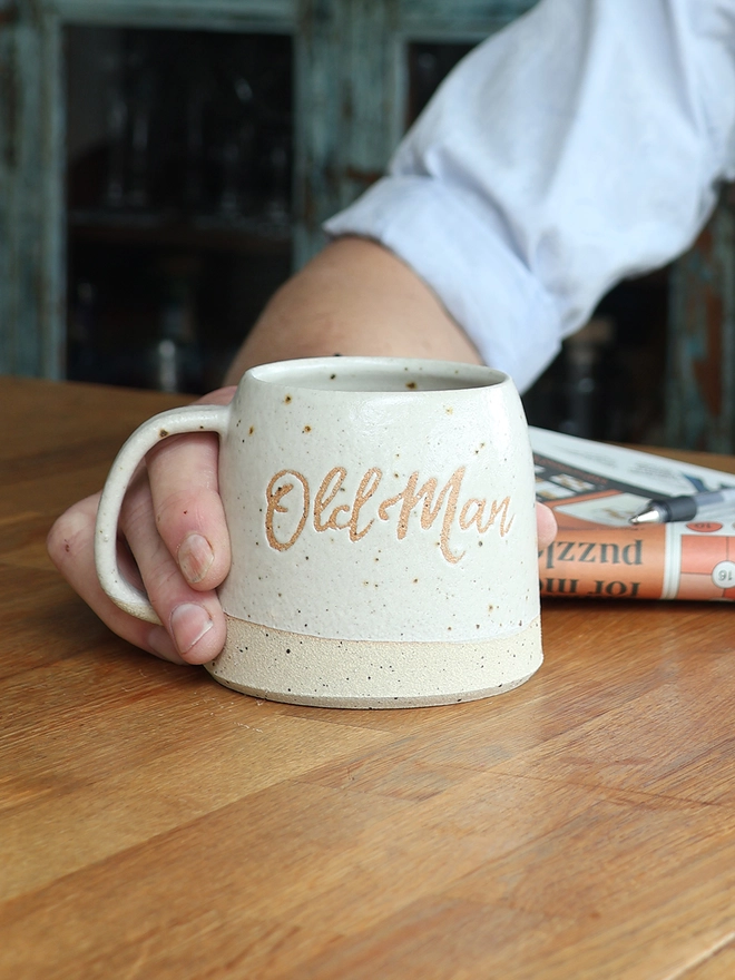 Personalised handmade ceramic stoneware mug featuring the nickname 'Old Man'. A hand holds the mug which is sat on a wooden table
