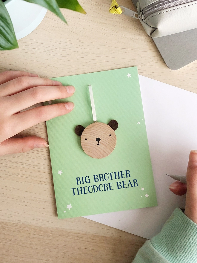 A green greetings card with a small wooden bear keepsake and the words "Big brother Theodore bear" printed on is on a wooden desk. 